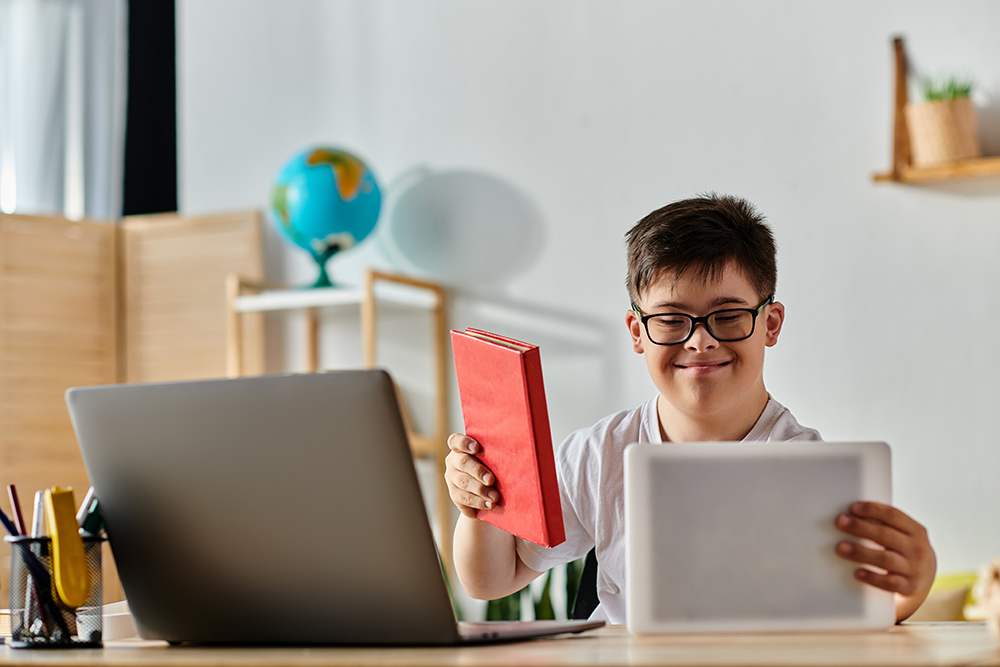 A boy with Down syndrome seated at a desk with a laptop and book.